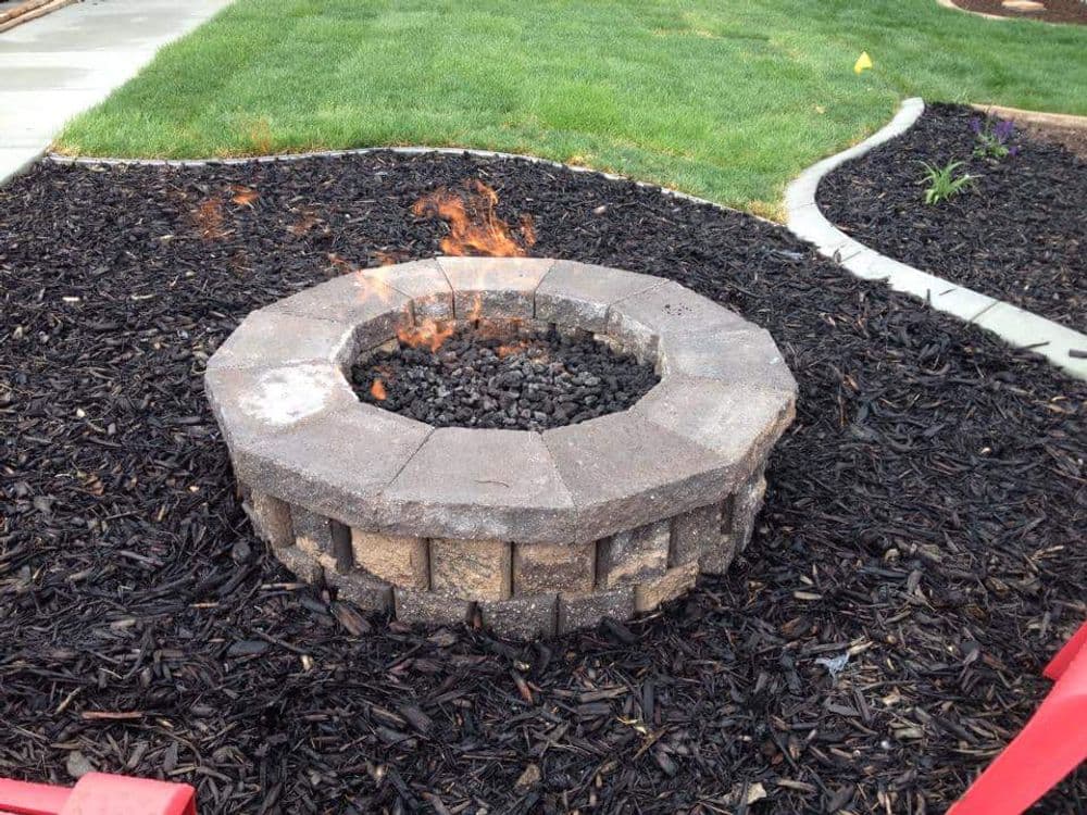 Outdoor stone fire pit with flames surrounded by black mulch and green grass.