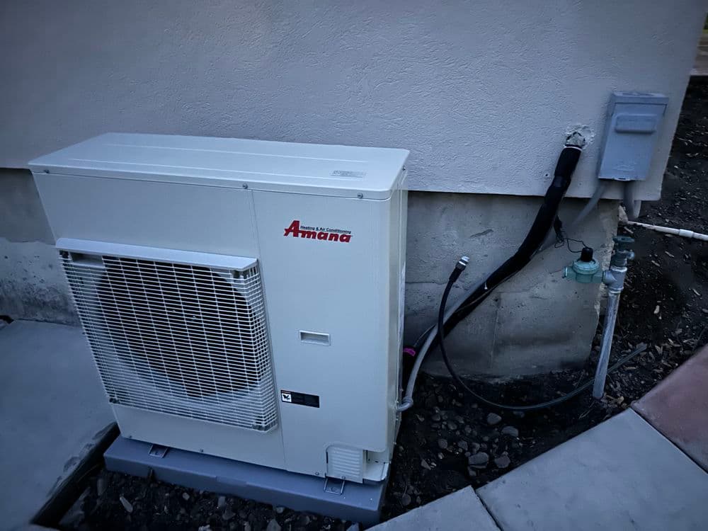 Amana air conditioning unit installed outside a building, featuring pipes and electric connections.