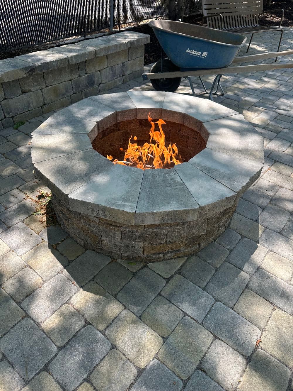 Outdoor stone fire pit with flames, surrounded by paver patio and a wheelbarrow nearby.