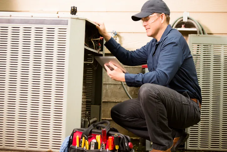 HVAC Replacement In West Jordan, UT 1 Man with a baseball cap crouching and working on an outdoor HVAC unit.