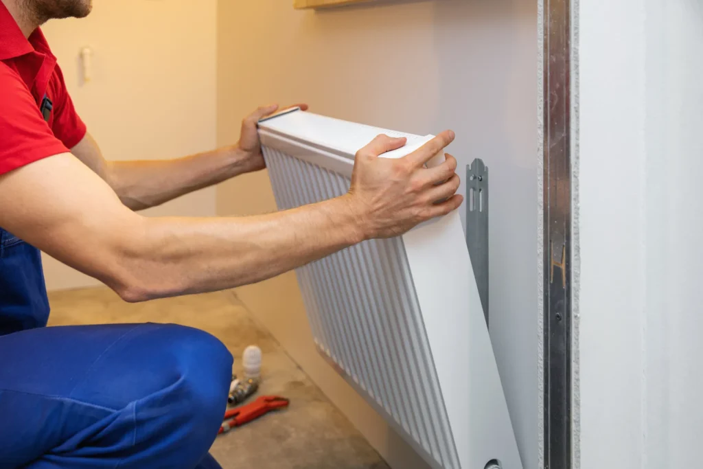 Heating Maintenance Service in Taylorsville 1 Technician in red shirt and blue pants installing white wall-mounted radiator using tools and fittings, aligning unit on metal brackets
