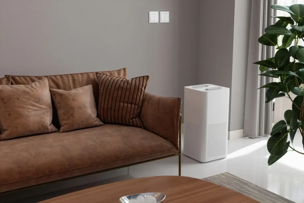 Whole Home Dehumidifiers in Sandy, UT 1 A white whole home air dehumidifier stands next to a brown velvet sofa with throw pillows in a modern living room, with a large green plant and a window with sheer curtains visible to the right.
