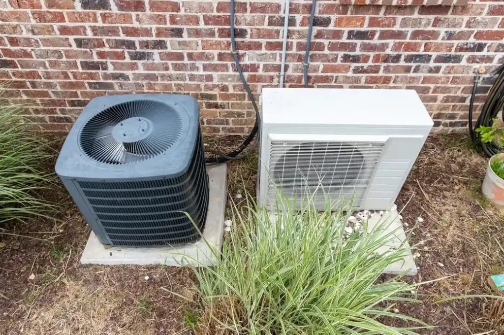 Mini Split AC Services in Taylorsville, UT 1 Two outdoor air conditioning units on concrete slabs beside brick wall—larger black unit with top fan and smaller white unit with protective grille, surrounded by grass and plants
