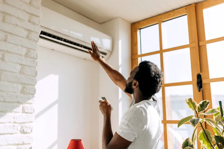 Mini Split AC System In West Jordan, UT 1 A person reaching up towards a wall-mounted mini-split air conditioner, holding a remote control.