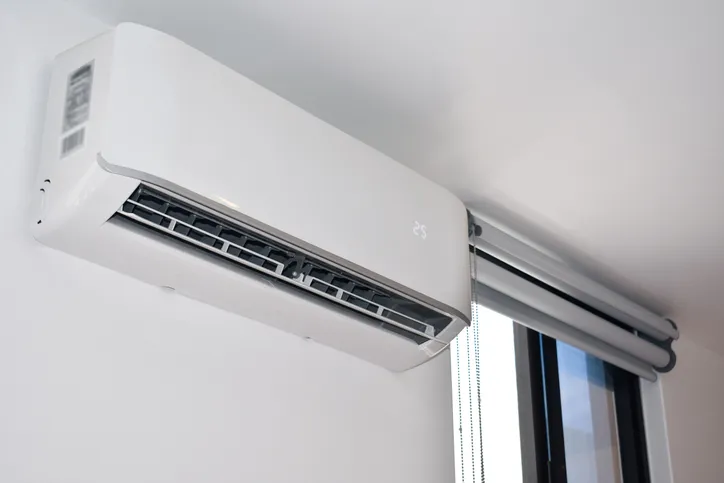 Mini Split AC Replacement In West Jordan, UT 1 A white mini-split air conditioner unit mounted on a white wall next to a window with blinds.