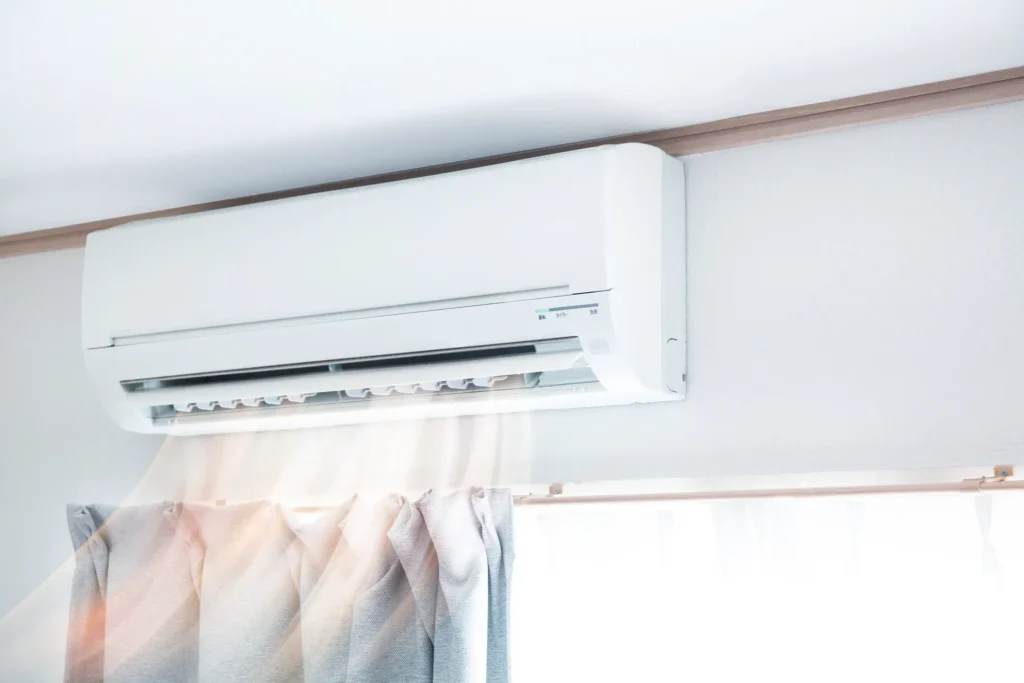 Wall-mounted air conditioning unit near ceiling actively blowing air above window with partially drawn curtains in naturally lit room